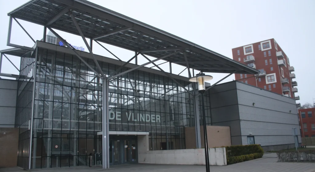 The glass entrance of "De Vlinder" building with modern steel architecture and nearby residential apartments.