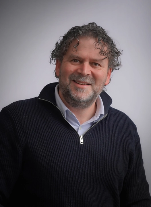 A man with curly hair and a beard wearing a dark sweater smiles against a neutral background.