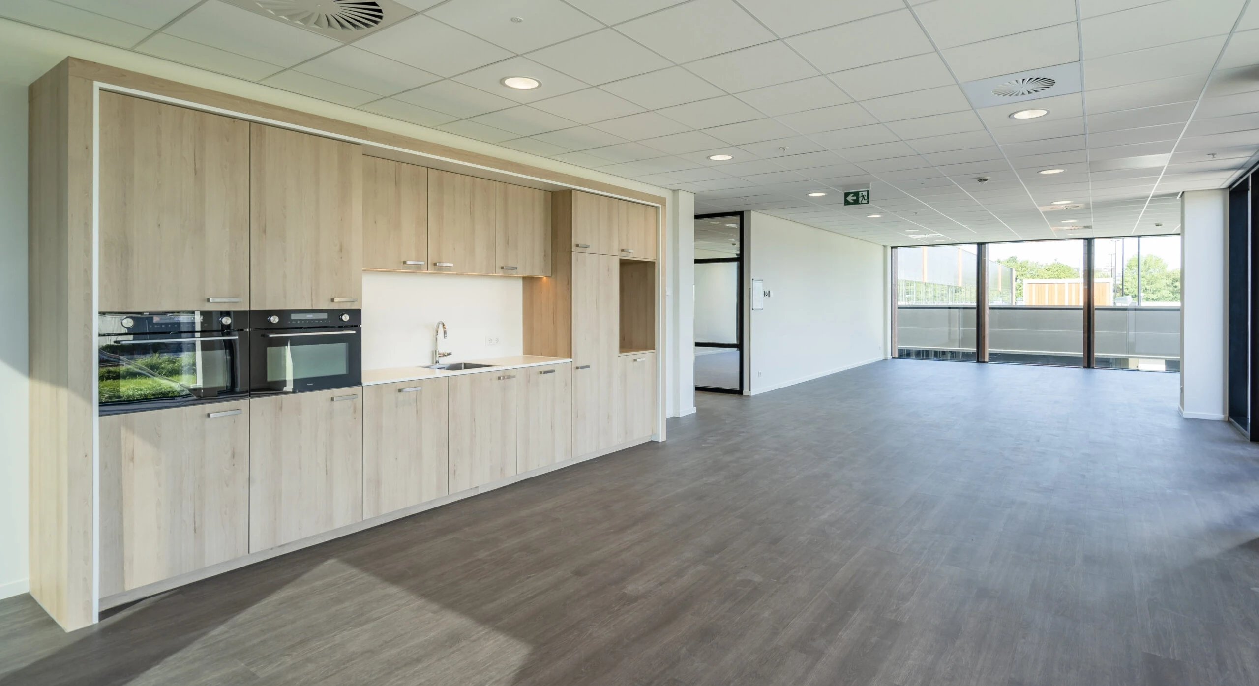 Modern kitchen space in an open-plan office area with wooden cabinets and large windows.