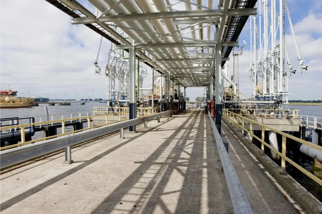 A pathway at an industrial refinery site adjacent to water, with pipes and equipment visible.
