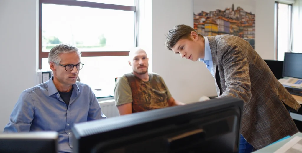 Three men are working together at a computer screen in an office.