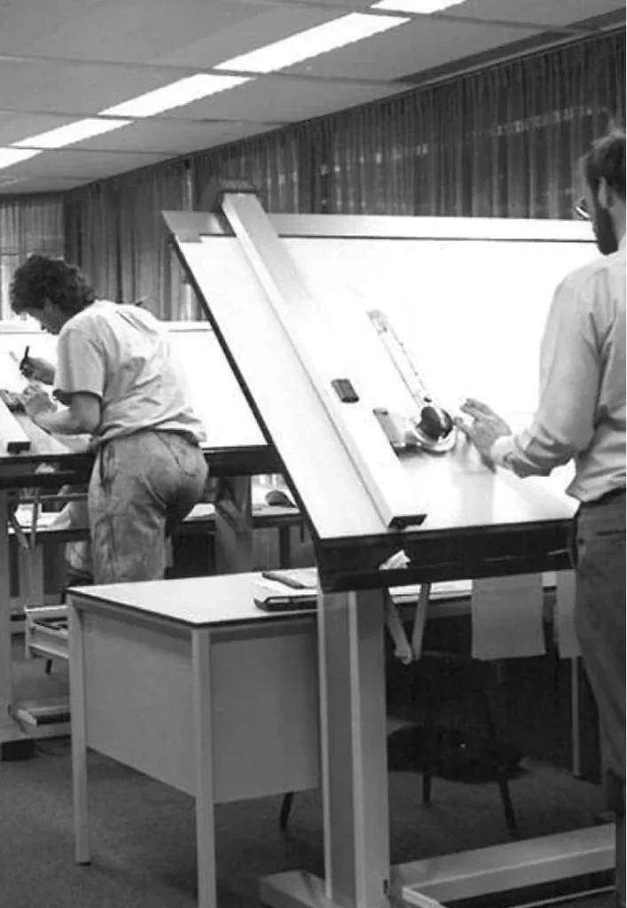 Two people are working at drafting tables in a vintage office setting.