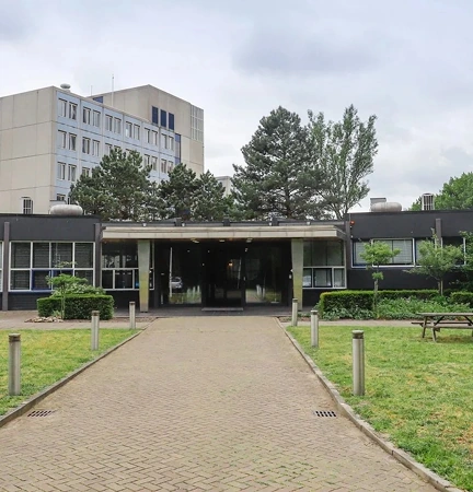 The image shows the entrance of a modern office building with surrounding greenery.