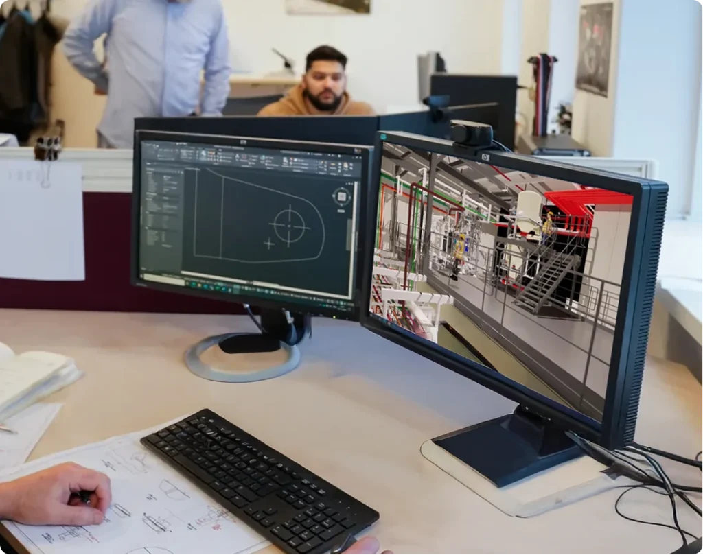 Two computer monitors displaying a 3D engineering drawing and a CAD design in an office setting with people working in the background.