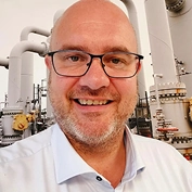 A smiling man wearing glasses stands in an industrial setting with large pipes and machinery in the background.