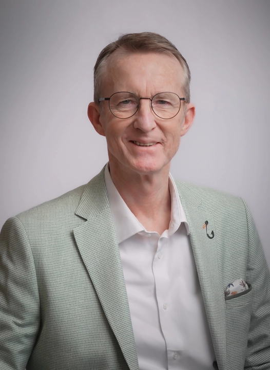 A man wearing glasses and a light green blazer smiles at the camera against a neutral background.