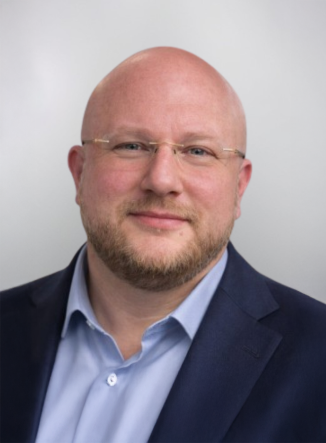 A bald man with glasses and a beard is wearing a suit and smiling at the camera.