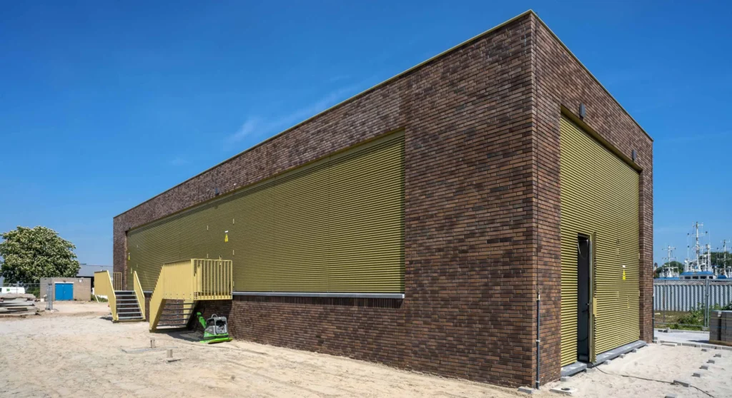 A modern industrial building with brown brick walls and yellow metal doors under a clear blue sky.