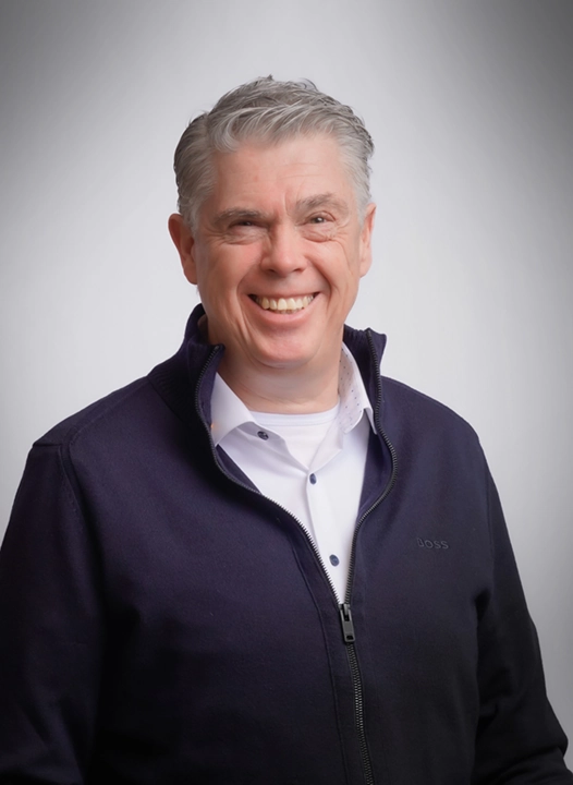 A smiling man with grey hair wearing a dark zip-up sweater over a white shirt.