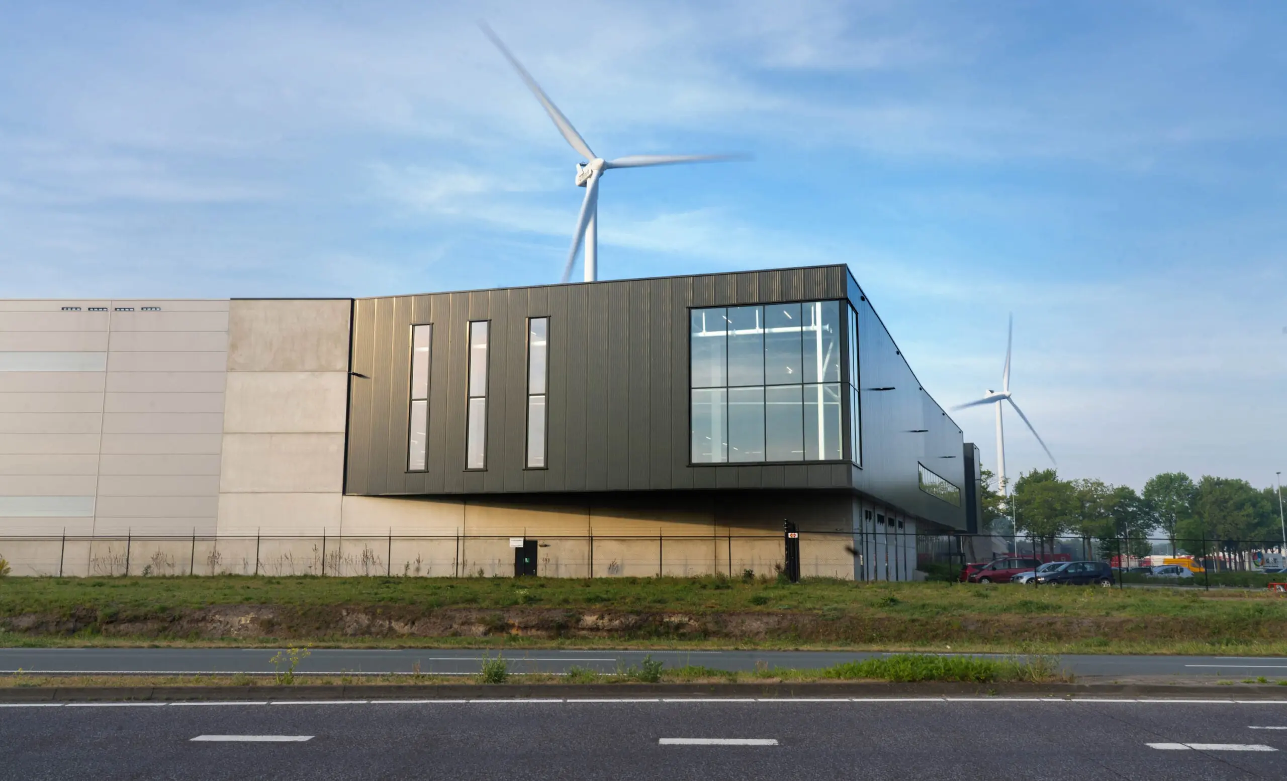 A modern industrial building with a wind turbine in the background under a blue sky.