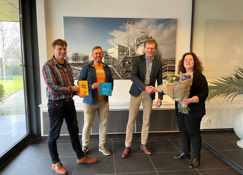 Four people are standing together, two holding books and one giving flowers.