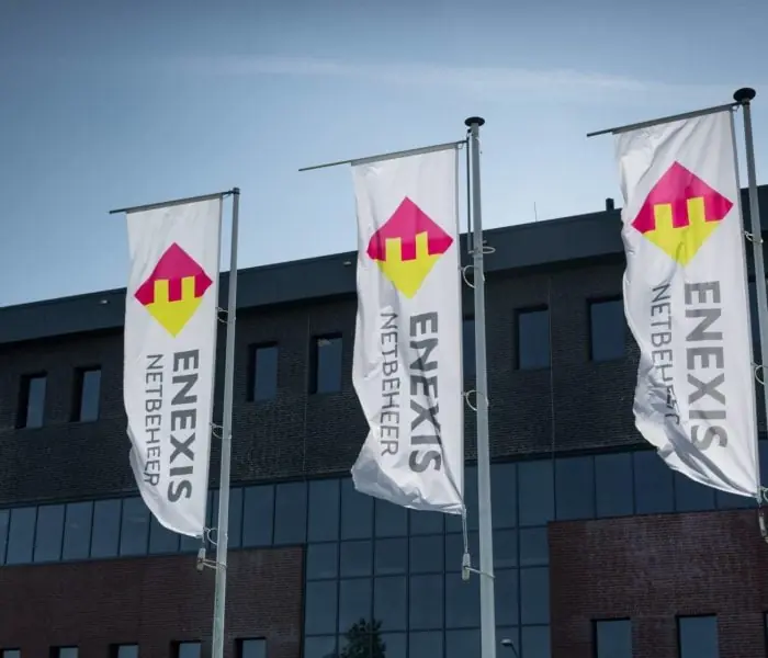 Three flags with the Enexis Netbeheer logo flying in front of a modern office building.